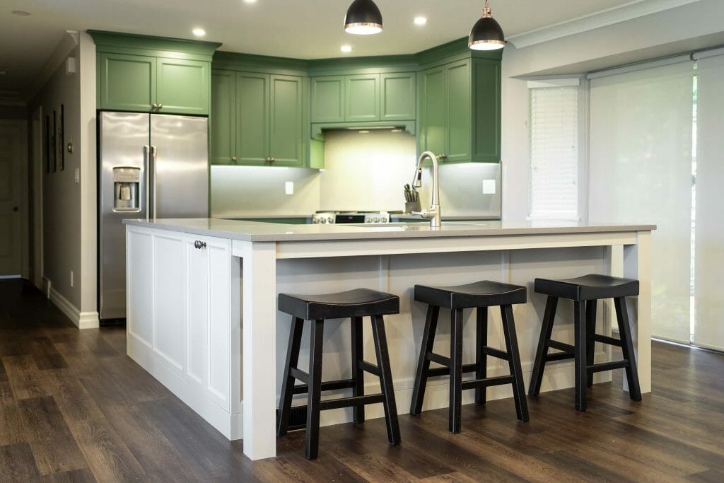 A renovated kitchen that has a large L-shaped island in white but green coloured kitchen cabinets.