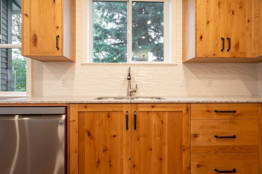 Kitchen cabinets in stained pine with sink, white tiled backsplash and a window above the sink.